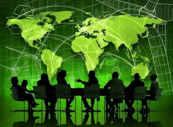 Painel eletrônico em cor verde mostra mapa-múndi, sobre ele imagens translúcidas de hélices eólicas, símbolos da reciclagem e números diversos, a frente do painel um grupo de pessoas se reúne e conversa a mesa.