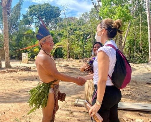 Em uma aldeia um homem de etnia indígena com cocar cumprimenta com um aperto de mão uma mulher branca de máscara.