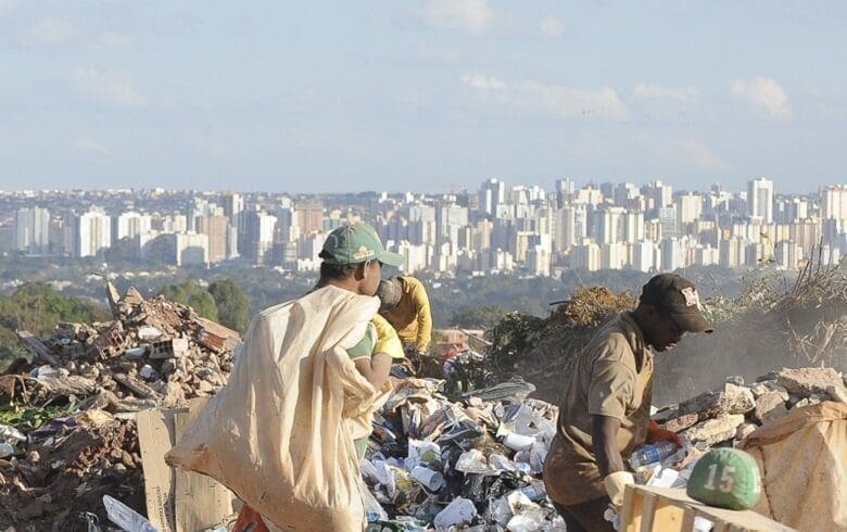 A imagem mostra um dos problemas sociais do Brasil, dois homens em meio à um lixão segurando sacolas e ao fundo da paisagem vários prédios.