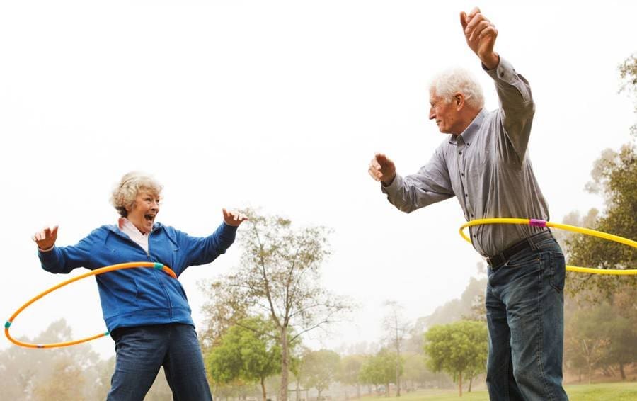 Envelhecimento saudável: dois idosos (uma mulher e um homem) brincando com um bambolê em um parque natural.