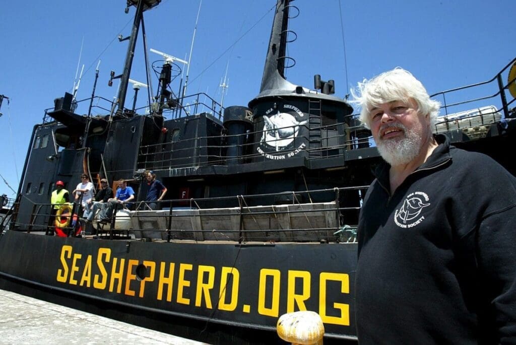 Homem de meia idade, cabelos e barba branca, aparece a frente de um navio preto com a inscrição "Sea Shepherd.org".