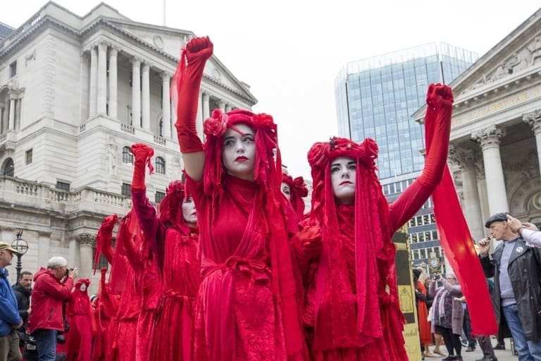 Extincition Rebellion -como surgiu esse movimento ambientalista. Mulheres vestidas de vermelho dos pés a cabeça e rosto pintado de branco, protestam em uma cidade com braços levantados e "mão em punho".