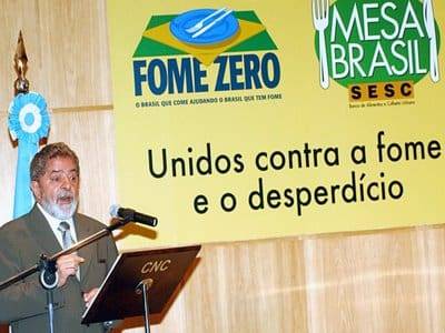Fotografia do Presidente Lula lançando o Programa Fome Zero em uma tribuna. Ao fundo o cartaz da campanha com a frase: "Unidos contra a fome e o desperdício".