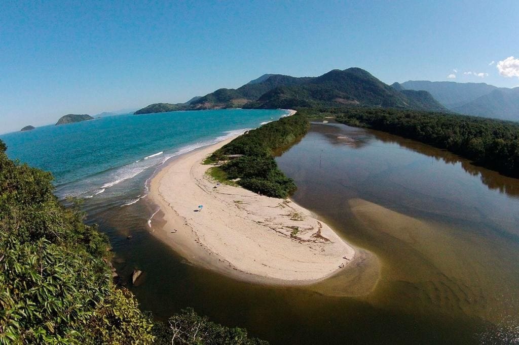 Vista de um estuário, onde as águas doces de um rio se encontram com as águas salgadas do mar. A paisagem mostra canais sinuosos, bancos de areia e áreas de vegetação costeira, como manguezais, que abrigam aves e outros animais. O ambiente apresenta transição suave entre terra e água, formando um ecossistema rico em nutrientes e biodiversidade.