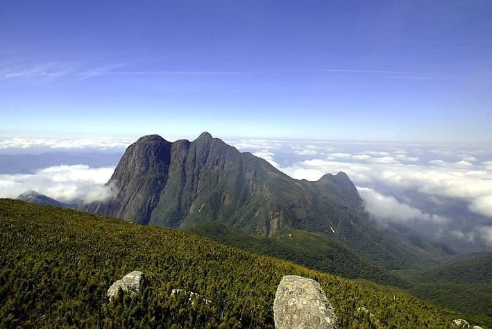 Parque Estadual Pico Paraná tem ao centro uma alta montanha rodeada por nuvens e céu azul