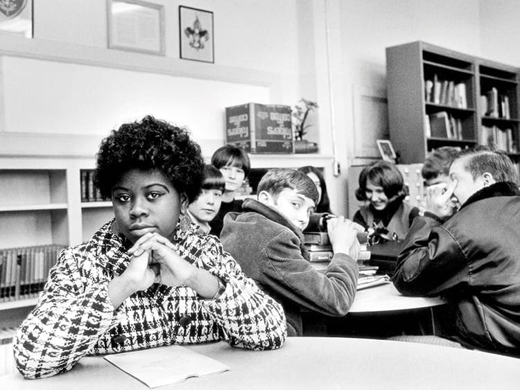 Fotografia ilustrando a segregação racial: uma negra sentada em uma biblioteca em uma mesa separada dos demais colegas brancos, que olham para ela de forma discriminatória.
