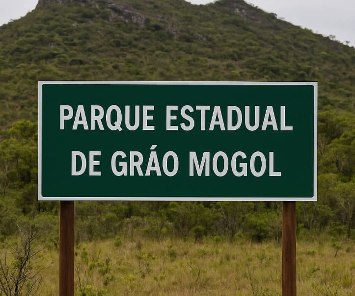Placa verde exibe: “Parque Estadual de Grão Mogol” ao centro de vegetação nativa e morros rochosos ao fundo.